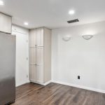 A kitchen with stainless steel refrigerator, white cabinets, wood flooring, and white walls. Two wall-mounted lights are on the right above an empty space.