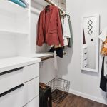A walk-in closet with white shelves, drawers, and hanging rods. Clothes, including a red blazer and a floral shirt, are hanging. A wire laundry basket sits on the wood floor, and a mirror is mounted on the wall.
