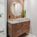 Modern bathroom with a wooden vanity, round mirror with backlighting, potted plant, and a glass-enclosed shower. The walls are white and the overall decor is minimalist.