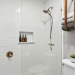 A modern white-tiled shower with glass doors, a brass showerhead and handle, three bottles in a built-in niche, and folded towels on a wooden shelf above a toilet with a small potted plant.