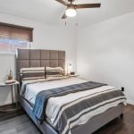 A modern bedroom with a gray upholstered bed, striped bedding, a side table with a plant and lamp, dark wood floors, white walls, and a ceiling fan. A window with a brown shade lets in natural light.
