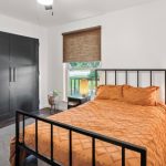 A bright bedroom with a black metal bed frame, orange bedding, two bedside tables with lamps, a large black closet, a window with a brown shade, and a plant on the floor.