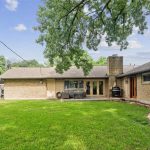 A spacious backyard with green grass, a large tree, a patio area with outdoor furniture and a grill, and a single-story brick house with several windows and doors under a blue sky with scattered clouds.