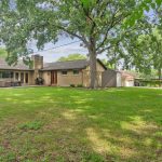 A spacious backyard with green grass, large mature trees, and a brick house featuring a patio area, outdoor grill, and shed. The scene is bright and peaceful with clear skies and scattered clouds.