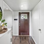 A bright entryway with white walls, a wooden front door, dark wood flooring, a round mirror, and a potted plant on a tray atop a white cabinet.