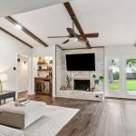 A bright living room with wood floors, white walls, exposed ceiling beams, a ceiling fan, a white brick fireplace, wall-mounted TV, built-in shelves, large windows, and glass doors opening to a green backyard.