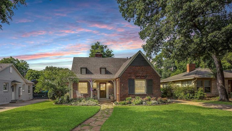 A charming brick house with a steep roof, large windows, and a front porch sits amid a well-kept lawn and garden, surrounded by mature trees, under a colorful evening sky.