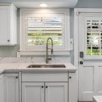 Bright kitchen with white cabinets, marble countertops, a stainless steel sink below a window with shutters, light blue tile backsplash, and a white door with a window leading outside.