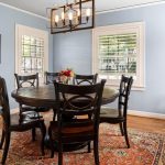 A dining room with light blue walls, a round dark wood table, six matching chairs, an ornate rug, a chandelier, two windows with shutters, and framed paintings on the walls. A vase of flowers sits on the table.