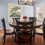 A dining room with a round dark wood table, six matching chairs, a red patterned rug, pale blue walls, two windows with white shutters, a flower centerpiece, and a framed painting on the wall.