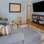 A cozy living room with a gray sectional sofa, striped pillows, a glass coffee table with a flower arrangement, a large wall-mounted TV, and a wooden media console on a patterned rug over hardwood floors.