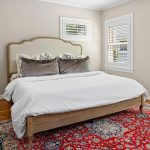 A cozy bedroom with a wooden bed frame, white bedding, decorative pillows, light-colored walls, wooden floor, large ornate red area rug, and two windows with white shutters.