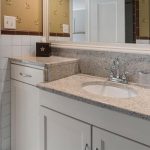 A bathroom vanity with a granite countertop, built-in sink, chrome faucet, white cabinets, and a large mirror. The wall has beige wallpaper with a floral pattern and a tile backsplash.