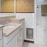 A bathroom with a granite countertop, white cabinets, a built-in sink, a large mirror, white tile walls, hexagonal floor tiles, a towel hanging on a rack, and a heater below shelves.