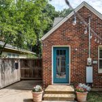 A small brick house with a blue door, string lights overhead, two flower pots by the entrance, and a wooden fence on the left. Large trees provide shade in the background.