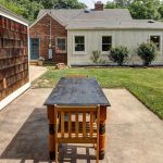 A wooden table and chairs sit on a concrete patio in a backyard with green grass, shrubs, and a house with brick and white siding in the background. A wooden-sided structure is on the left.