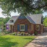 A charming brick house with a steep roof, large front window with dark shutters, manicured lawn, garden beds, a tree-shaded driveway, and a purple flag hanging by the front porch.