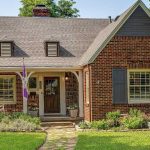 A charming brick house with a steep gabled roof, dormer windows, a covered porch with a rocking chair, dark shutters, and a well-kept front yard with flowers and shrubs.
