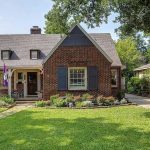 A charming brick house with a gabled roof, front porch, and shuttered windows sits amid a well-kept lawn and landscaped garden, with a driveway on the right and trees and neighboring house in the background.