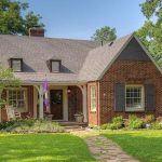 A charming brick house with a steep gabled roof, rocking chair on the porch, purple flag by the door, green lawn, flowerbeds, and trees surrounding the home on a sunny day.