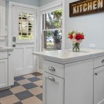 A bright kitchen with white cabinets, checkered tile floor, a vase of flowers on a white island, a window and glass door letting in sunlight, and a “Kitchen” sign above the door.