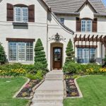 A two-story house with cream-colored brick, dark wooden shutters, and a brown front door. Manicured shrubs and flower beds line the pathway leading to the entrance, with a green lawn and blue sky in the background.