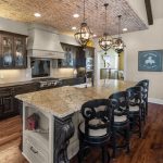 A spacious kitchen with dark wood cabinets, a large granite island with three black chairs, hanging pendant lights, and a brick ceiling. The floor is wooden, and there's a chalkboard on the wall in the background.