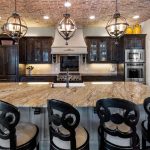 A spacious kitchen with a large granite island, five ornate black chairs, dark wood cabinets, stainless steel appliances, and two globe pendant lights hanging from a brick ceiling.