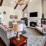 A cozy living room with vaulted ceilings, exposed wooden beams, and a fireplace. There are sofas, a coffee table, a TV above the fireplace, and large windows letting in natural light.