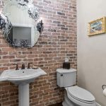 A bathroom with exposed brick walls, a white pedestal sink, ornate mirror, white toilet, dark wood floor, wall-mounted light fixtures, a chandelier, and a framed painting on a beige wall.