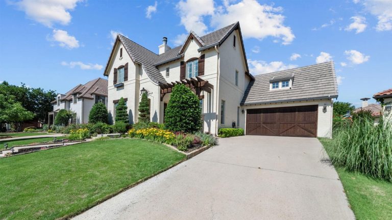A two-story white brick house with a brown tile roof, wooden garage door, manicured lawn, and colorful flowerbeds under a blue sky with scattered clouds.