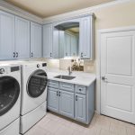 A modern laundry room with light blue cabinets, a white counter with a sink, a front-loading washing machine and dryer, tiled floor, and a white door. There is also a small cushioned bench on the right side.