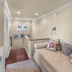 A cozy upstairs nook with a daybed adorned with floral and patterned pillows, soft lighting, framed photos on the wall, wood flooring, and a TV on a white cabinet. A staircase with a wooden handrail is visible.