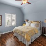 A cozy bedroom with blue walls, hardwood floor, a bed with yellow and white bedding, two side tables with lamps, a patterned armchair, a framed poster, and two windows with white shutters.