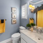 A bathroom with blue walls, a white sink with a marble countertop, a toilet, yellow towels on a rack, butterfly wall art, a mirror, plants, and a yellow shower curtain. Sconces with yellow shades are above the sink.
