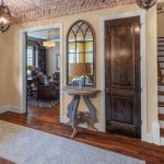 A warm entryway with a wooden door, arched brick ceiling, a light blue rug, a small decorative table with a lamp and mirror, dark wooden doors, and stairs with white risers and wood treads.