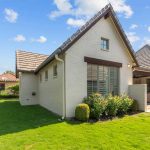 A modern house with white brick exterior, sloped roof, and large windows faces a well-kept green lawn and landscaped bushes under a bright blue sky with scattered clouds. Outdoor seating is visible on the patio.