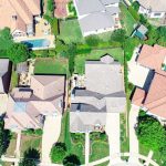 Aerial view of suburban homes with driveways, green lawns, trees, fences, and backyard swimming pools on a sunny day.