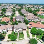 Aerial view of a suburban neighborhood with rows of houses, green lawns, trees, and curved streets under a clear blue sky.