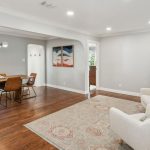 Modern living and dining room with hardwood floors, neutral walls, two armchairs and a patterned rug in the foreground, a wooden dining table with six chairs, large windows, and contemporary art on the wall.
