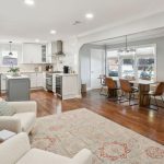 Open-concept living space with a cozy sitting area, a light gray kitchen featuring stainless steel appliances, and a dining area with a wooden table and six chairs by large windows, all with hardwood floors.