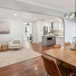 Open-concept dining and living area with a wooden dining table and leather chairs, light armchairs, a rug, and a modern kitchen in the background. Walls are light-colored, creating a bright and inviting space.