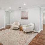 A bright living room with two cream armchairs on a patterned rug, a small round wicker table, light gray walls, wood flooring, and an archway leading to a sunlit adjoining room.