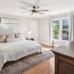 A bright, modern bedroom with a large bed, neutral bedding, matching nightstands with lamps, a ceiling fan, a window with white curtains, and a wooden dresser. A decorative sign hangs above the bed.