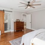 A bright bedroom with hardwood floors, a bed with white bedding, a wooden dresser, a leaning mirror, and a sliding barn door leading to a bathroom. A ceiling fan and rug complete the cozy decor.