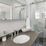 Modern bathroom with a gray countertop, sink, soap dispenser, towel basket, large wall mirror, and a glass shower with marble-patterned tiles and hexagonal floor tiles.