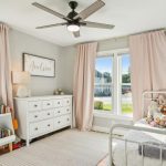A bright, tidy child's bedroom with light gray walls, pink curtains, a white dresser, bookshelves filled with books, stuffed animals on a white bed, and large windows letting in natural light.