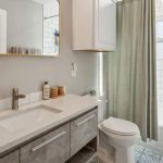 Modern bathroom with a gray vanity, white countertop, rectangular mirror, and white subway tile walls. There is a toilet, a bathtub with a green curtain, and various toiletries on the counter and in the shower area.
