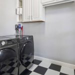 A laundry room with black Samsung washer and dryer, white cabinets above, a checkered black and white tile floor, and cleaning supplies stored in the corner.