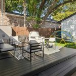 A modern backyard with a raised deck featuring outdoor seating, a small table, and cushions. The yard has artificial grass, a geometric climbing dome, string lights, and is surrounded by a wooden fence and trees.